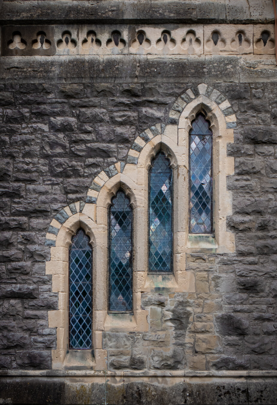 Four stepped lancet windows in a dark stone church wall. The sharply arched windows, stepping up from left to right, have diamond patterned leaded glass with the sun illuminating the odd pane. The windows are framed by a lighter, tan coloured stone alternating with darker stone over the top of the arches for a banded effect. A decorative border with carved trefoil arches runs along the top of the image.