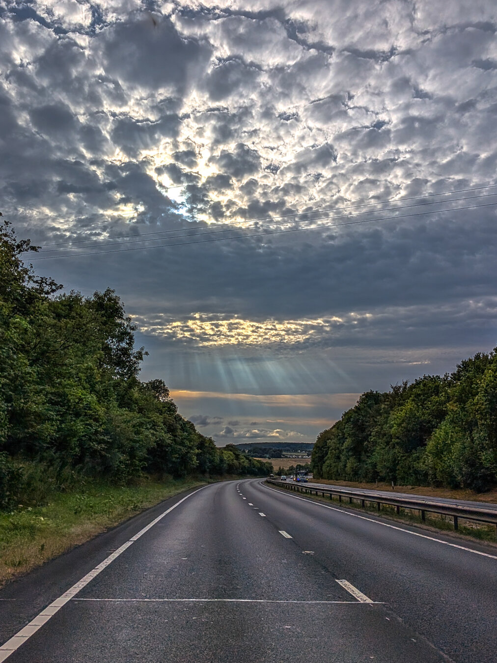 A long, curving, wide open highway with lush green trees bordering both sides. Overhead the sky is filled with a thick layer of heavily textured clouds. A break in the clouds ahead lets sunbeams break through and lights the horizon with pastel coral and blue.