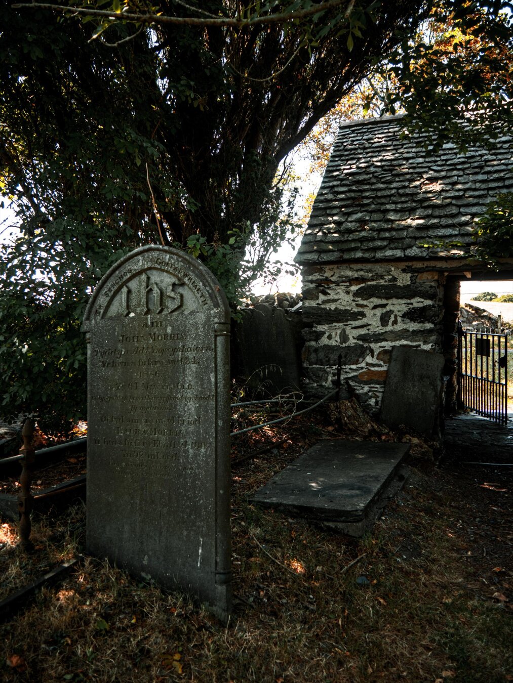 A weathered 19th-century slate tombstone dedicated to John Morris features elegant carvings and an "IHS" christogram monogram. It stands under a dense tree canopy beside a rustic stone lychgate with a wrought-iron gate.