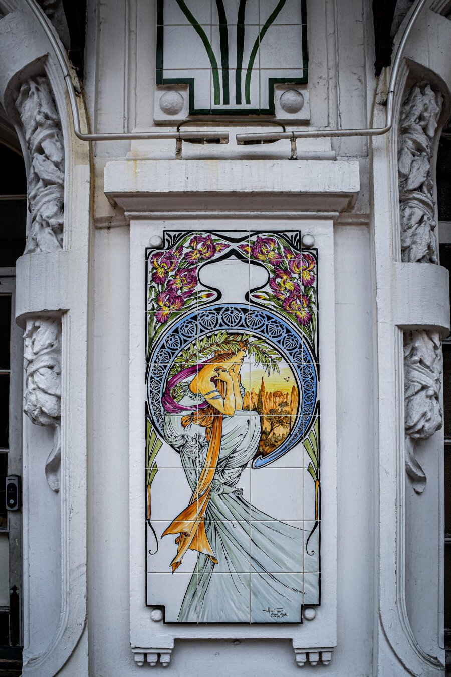 An Art Nouveau ceramic tile panel of  Alphonse Mucha's "Poetry", featuring an elegant woman crowned with laurel leaves gazing over a sunset landscape, framed by a curved blue border and purple irises on a white exterior wall.