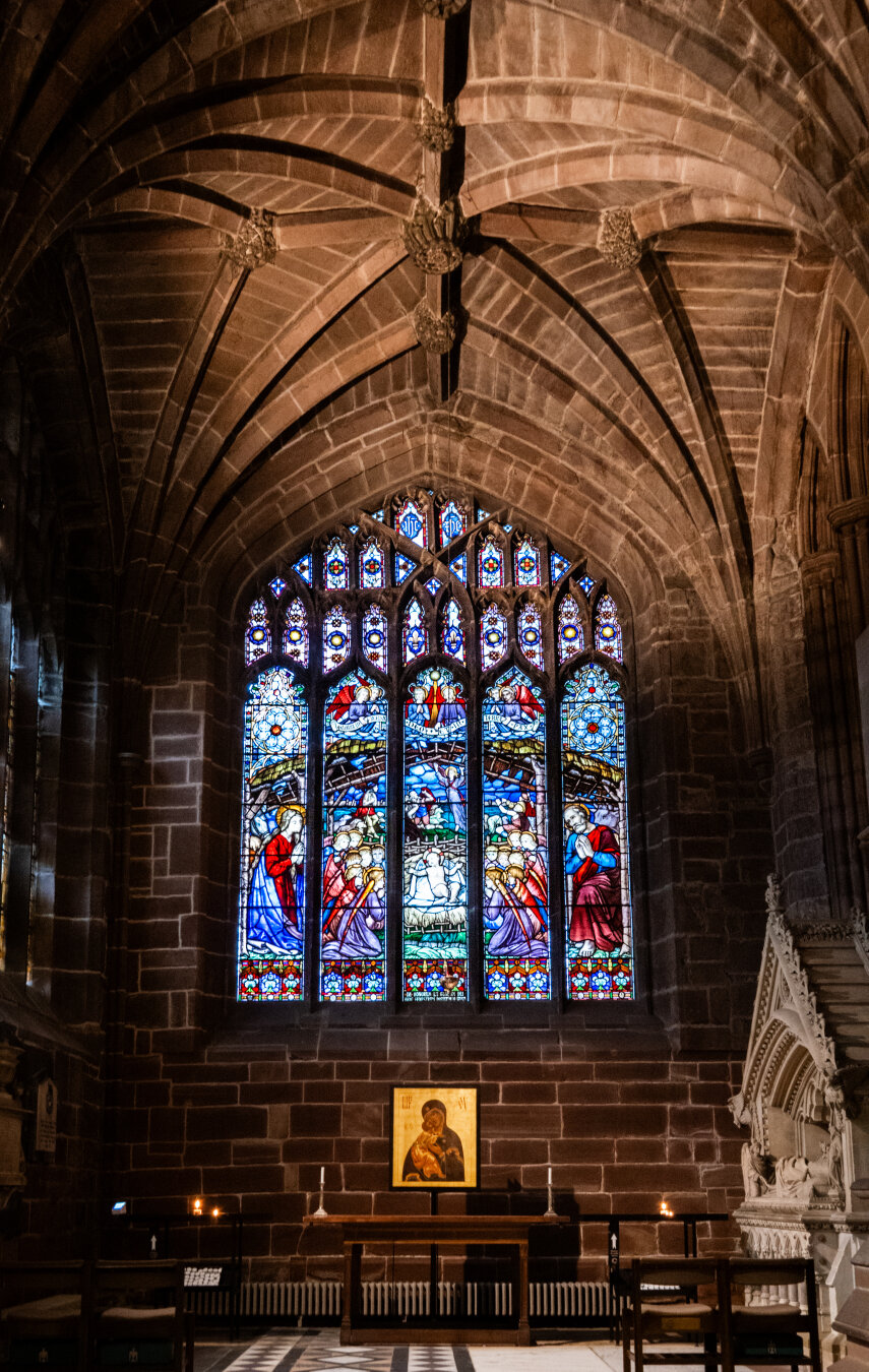 A vibrant stained-glass window depicting the Nativity scene inside Chester Cathedral. The Gothic architecture features dark stone block walls and dramatic pointed ceiling vaults.  A small altar table below the window holds candles and a "Madonna and Child" image.