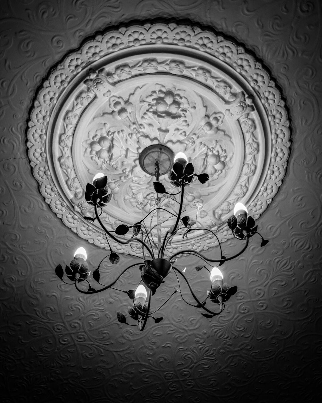 A high-contrast, monochrome, upward-facing shot of an ornate, wrought iron chandelier with glowing bulbs hanging from the center of a highly detailed Victorian plaster ceiling rose. The ceiling around it is embossed with intricate damask-patterned wallpaper.