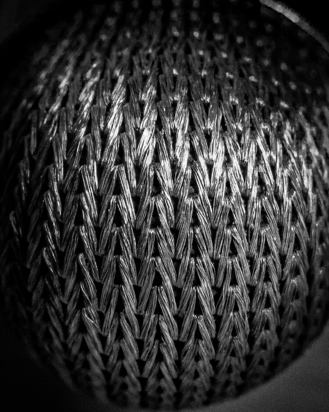 Monochrome, macro abstract photograph of a textured, silver-colored woven mesh with a tight herringbone pattern.