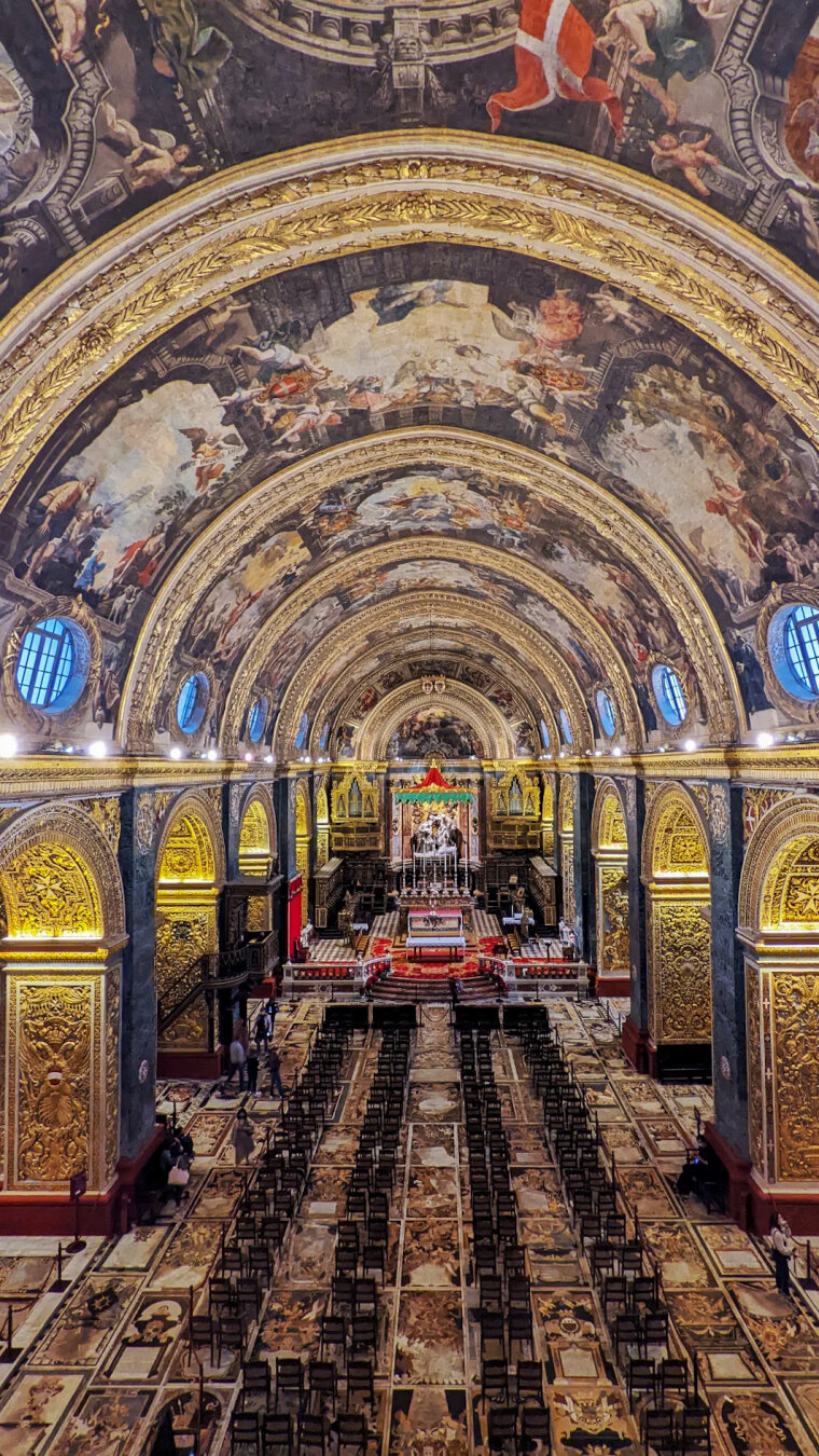 A high, wide-angle photo looking down the grand nave of St. John's Co-Cathedral in Malta. The vaulted ceiling is intricately painted and lead to gold, gilded side arches. Rows of chairs set on the marble floor below lead toward an illuminated high altar.