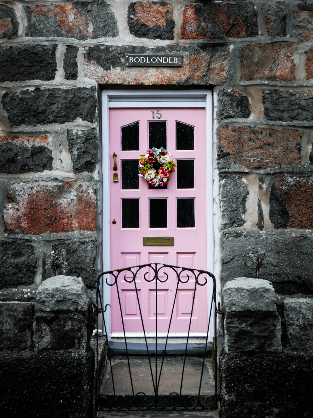 A light pink wooden front door with glass panels set in a charming stone cottage. A small, colorful flower wreath hangs on the front and a small slate sign with the house name "BODLONDEB" hangs over the door. In the foreground is a small iron garden gate.