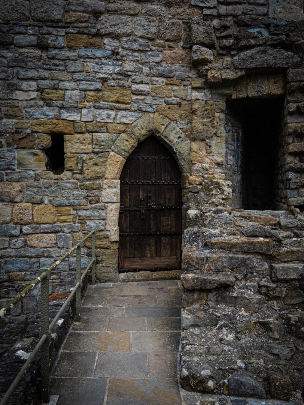 A heavy, dark arched wooden door with iron studs set in a rugged stone wall inside Caernarfon Castle.  A dark, narrow passageway sits to the right and a small arrow slit sits in the wall to the left.