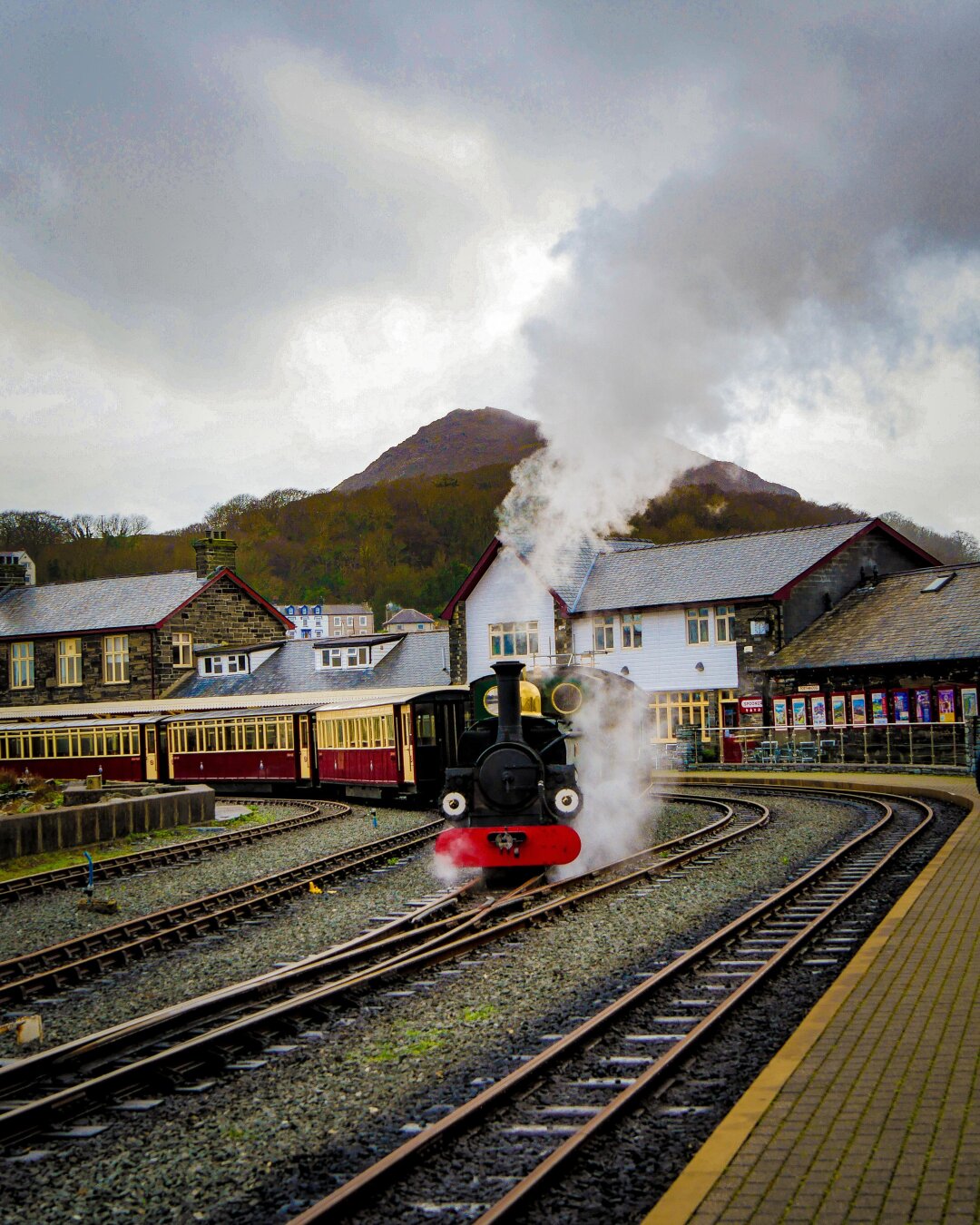 A narrow-gauge steam engine pulling a passenger train around a bend in a quaint station. The engine is directly facing the camera with two white headcode discs and a bright red bumper that look like a happy face. Thick white steam billows from the smokestack. Stone station buildings and a tree-covered mountain are visible in the background under grey clouds.