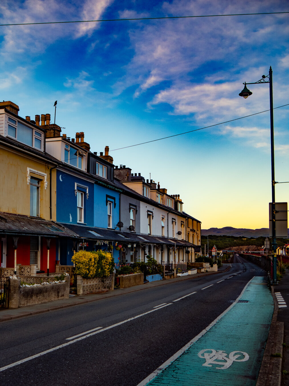 A row of brightly painted Victorian terraced houses in yellow and red, royal blue, sky blue, and white. A green bicycle lane runs along the right side of the dark asphalt road in front of the houses. A classic black street lamp stands on the right reaching into the dusk sky which is bright blue with wispy clouds and turns to yellow at the horizon.