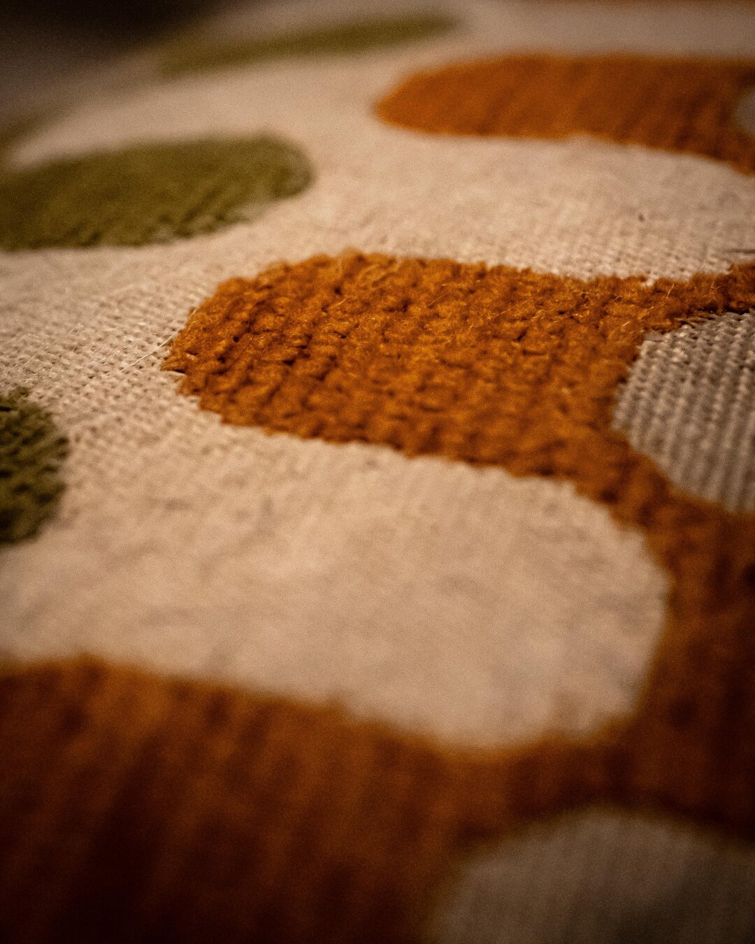 A macro close-up of a woven textile featuring abstract, organic shapes. The pattern includes textured loops of burnt orange and olive green fabric raised against a coarse, off-white background.