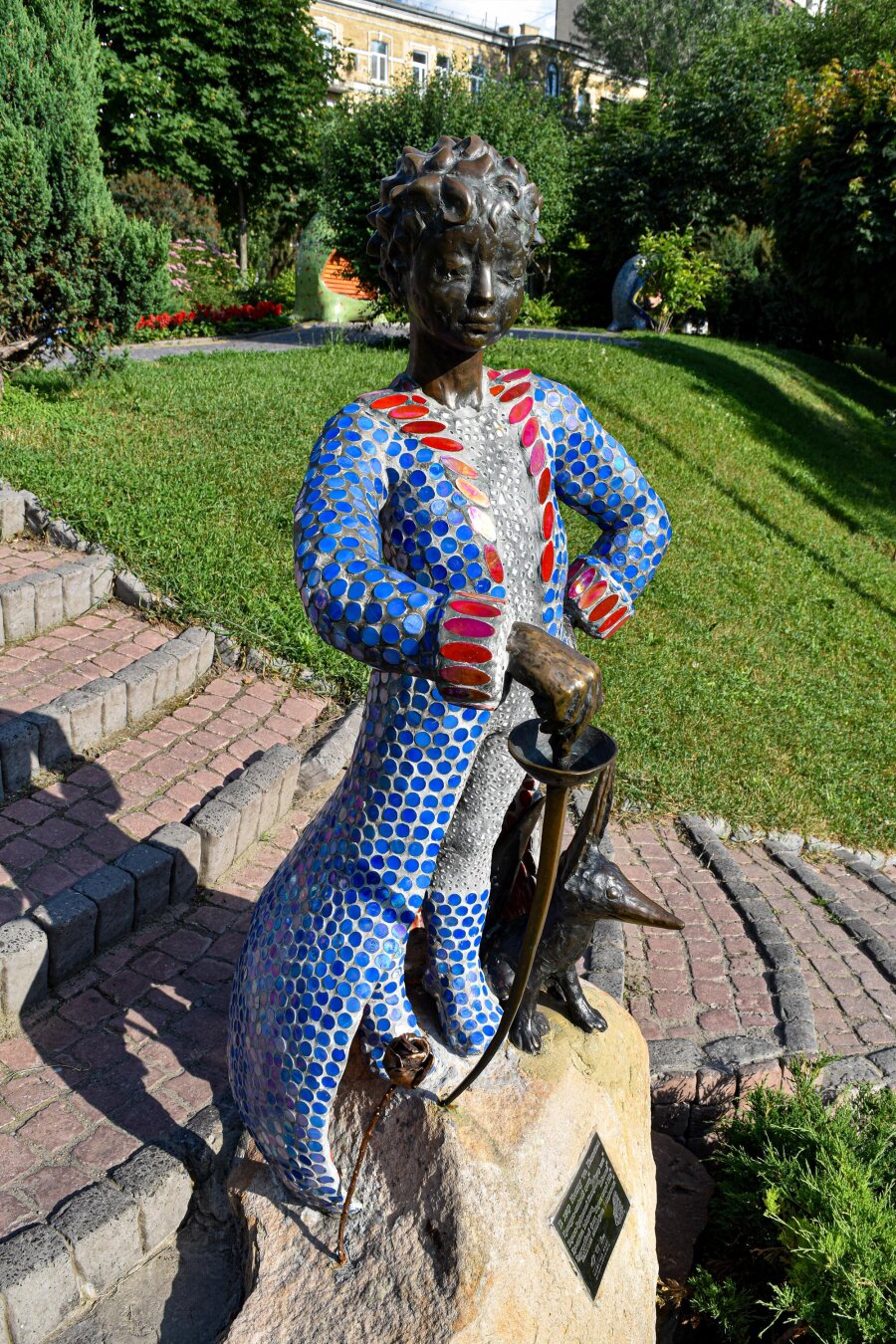 A bronze sculpture of the Little Prince wearing his long coat covered in blue and red mosaic tiles. He stands on a large stone pedestal in a park with a small bronze fox by his leg and a rose at his feet.