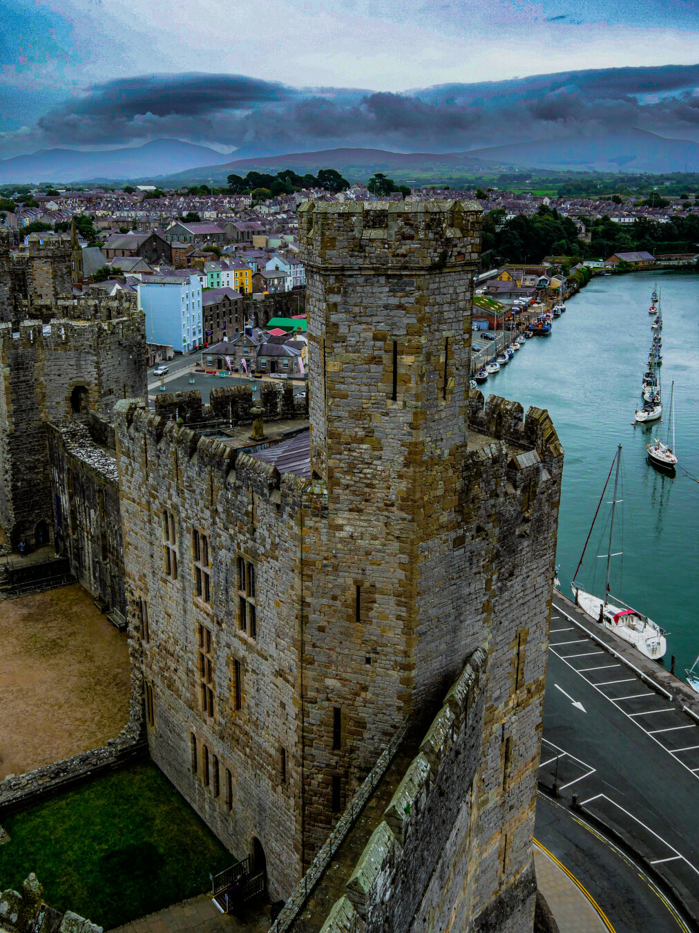A high-angle shot looking down at the massive stone towers and ramparts and the grassy courtyard of Caernarfon Castle. Outside the castle walls, to the right, a line of sailboats is moored in a calm river. Straight ahead in the background, a colorful Welsh waterfront town leads to rolling green hills under an overcast sky with low hanging clouds.