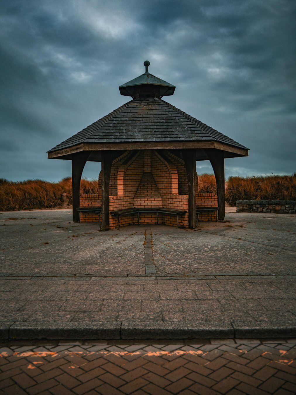 A symmetrical, octagonal gazebo stands on a paved beachfront promenade in Wales. The centre of the gazebo is made of brick with windbreaker walls between wooden benches. It is topped with a tiered, slate-shingled roof which is supported by wooden pillars that reach to the ground. In the background are tall amber dune grasses under a moody, overcast sky.