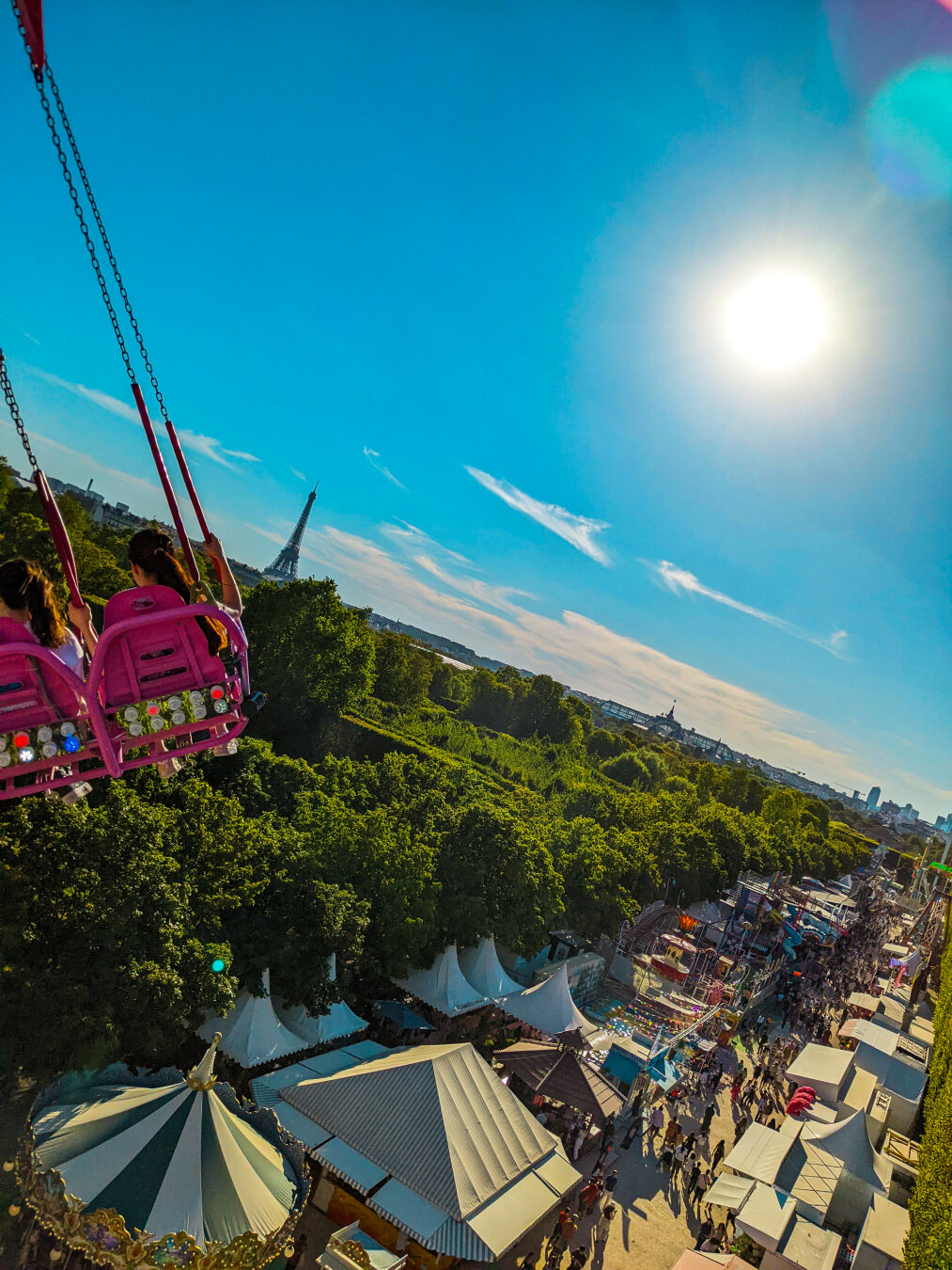 A tilted, wide angle aerial view from a swing ride at a fun fair in Paris. Two people with ponytail sit in bright pink plastic seats high over the fairground below. On the ground, the green and white top of a carousel and an array of white tents are set up among the green trees in the Jardin des Tuileries. In the distance, past the pink seats, the Eiffel Tower stands on the horizon under a vibrant blue sky with a bright sun.