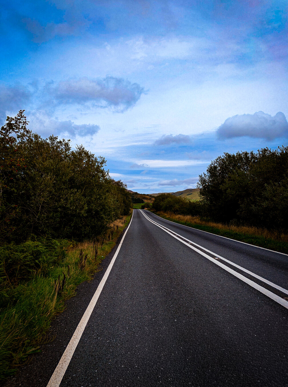 An open, empty road with white lane markings leads towards distant hills, with green trees and dense bushes on both sides under a cloudy blue sky.