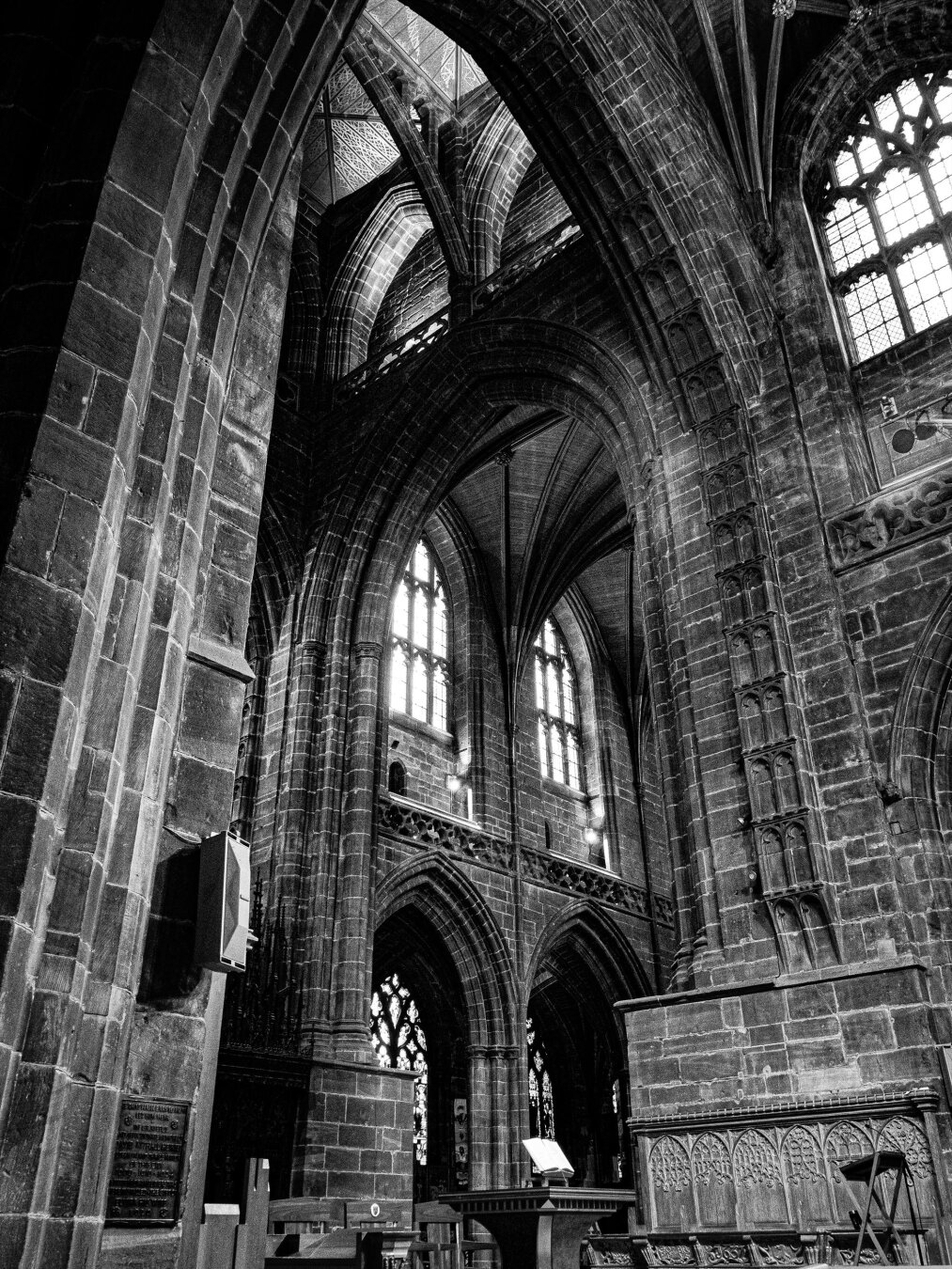 A high-contrast monochrome view looking up at the imposing interior of Chester Cathedral. The Gothic architecture features soaring stone arches, an intricately vaulted ceiling, and tall arched windows letting light into the magnificent space.