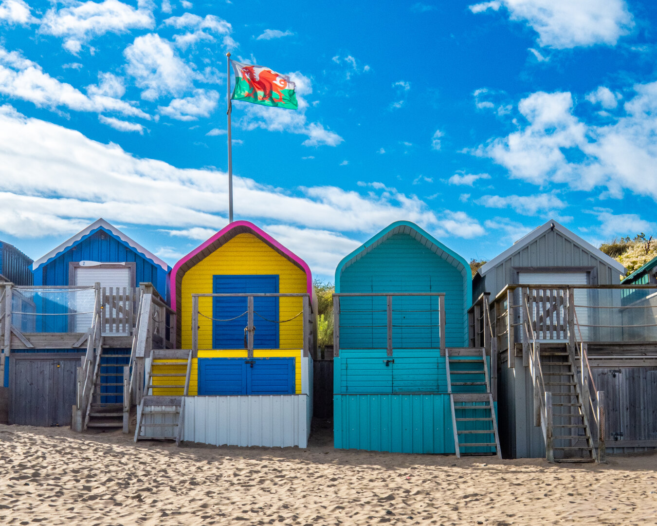 A row of four coastal beach huts standing on dry sand. From left to right, the huts are painted royal blue with white doors, bright yellow with blue doors and a pink roofline, solid teal blue, and light grey with a silver metal door. A tall flagpole stands behind the second hut, flying Welsh flag. The flag, known as Y Ddraig Goch (The Red Dragon) is white on the top half and green on the bottom with a red dragon centered in the middle.