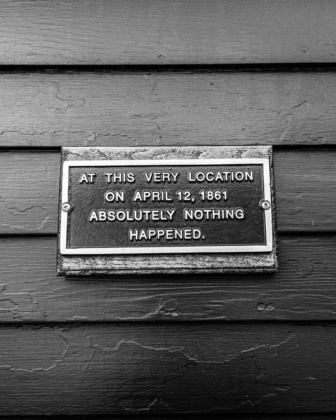 A black metal plaque on a wooden shingled wall. The plaque has a silver border and lettering that reads "AT THIS VERY LOCATION ON APRIL 12, 1861 ABSOLUTELY NOTHING HAPPENED."