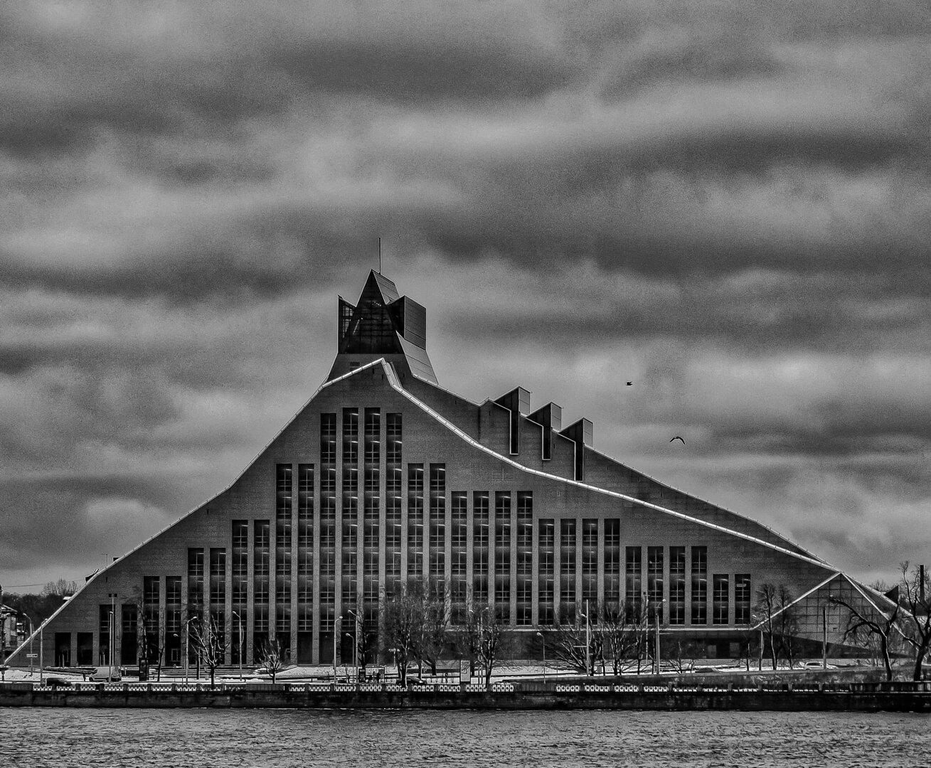 A monochrome view of the National Library of Latvia across the Daugava River. The building has an asymmetric, sloped facade which is lined with row upon row of rectangular windows that lead up to a peak with a three spoked crown on top. Bare winter trees line the water in front of the building under a moody, overcast sky.