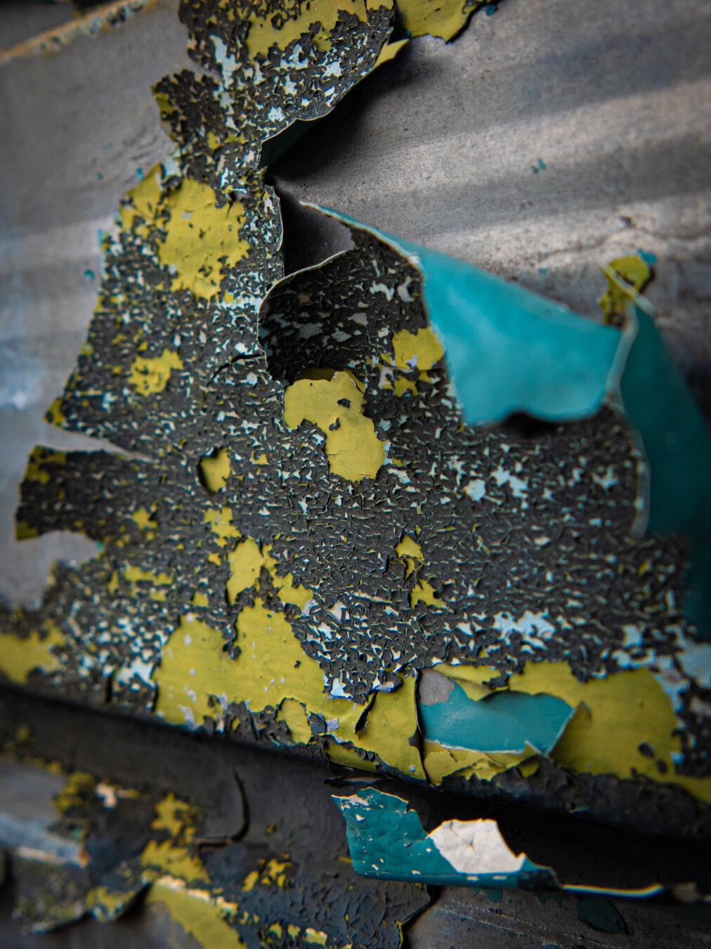Macro photograph of weathered industrial metal featuring layers of heavily cracked and peeling paint. A brown layer of paint is cracking to reveal a textured chartreuse yellow layer underneath which, in turn, is chipped to reveal a layer of teal paint at the bottom that is also curling up completely off the metal.