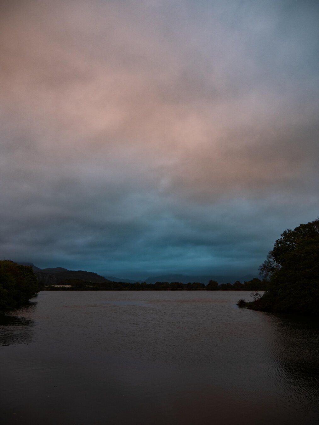 A dark lake at dusk surrounded by silhouetted trees. A heavy, overcast, dramatic sky fills the entire upper half of the image. Dark storm clouds hang over mountains on the horizon and soft pinkish-orange light comes through the clouds above.