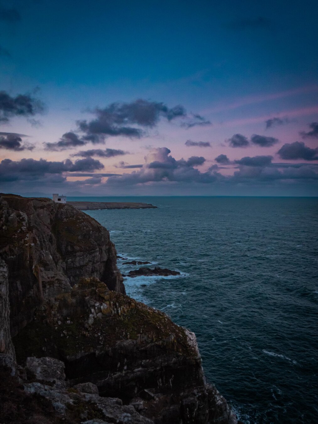 Coastal cliffs in Ynys Môn (Anglesey) at sunrise. A steep, rocky cliffside drops into a rough blue sea. At the top of the cliff a small white disused fog station can be seen. The sky is coloured pastel pink, purple, and blue with dark clouds scattered across it.