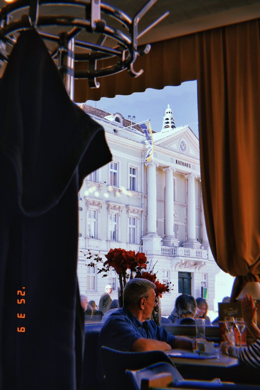 A crowded coffee place with reteo interior. Clothes rail in the foreground and the cities townhall visible through the big picture window in the background.