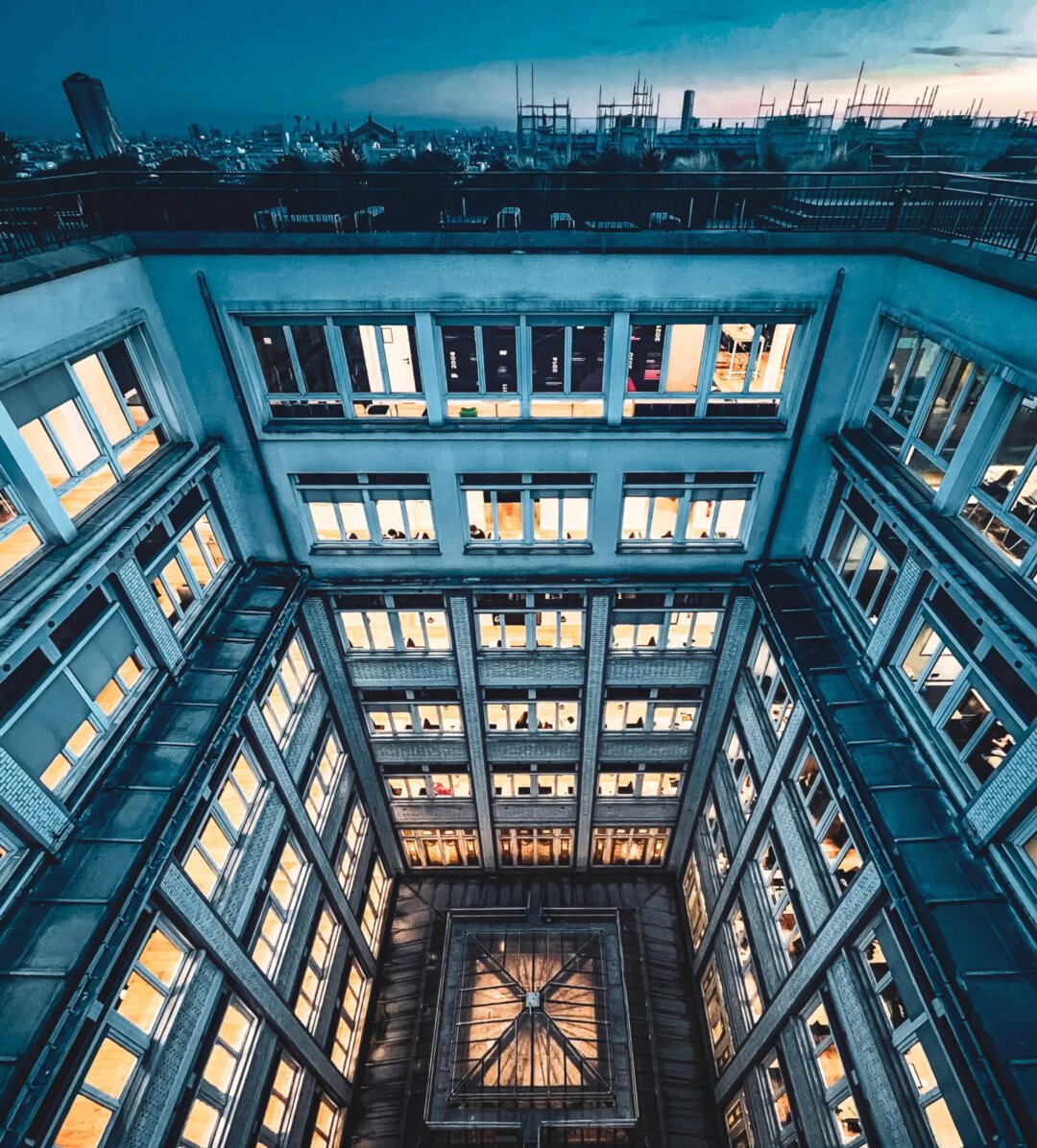 Photo prise depuis le toit d'un bâtiment. C'est la fin de journée. Le bâtiment est éclairé de l'intérieur. La photo a des teintes bleues et jaunes.