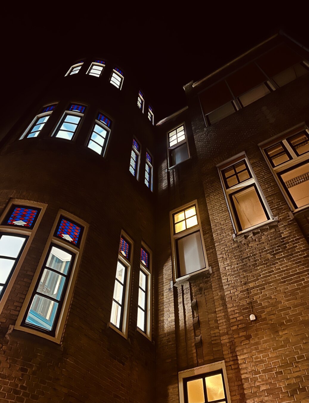 A photo of the front of an university building in amsterdam where two buildings collide exact in the middle of the photograph. The left side of the photograph has a cooler light beaming from the windows and beautiful mosaic windows. The right side has a warmer light shining within. Both contrasting  iconically!