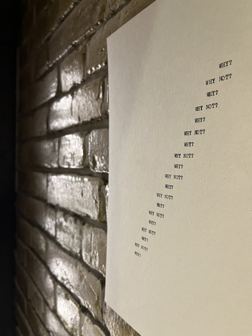 A photo of a concrete poem hanging on the wall; the poem repeats; why? why not? Why why not? Why? why not? All the way from the bottom left to the top right of the page.