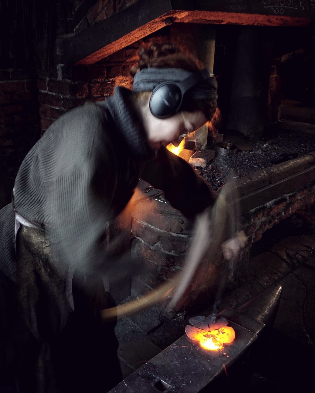 Photo of a woman in grey woolen sweater, striking glowing iron with a hammer