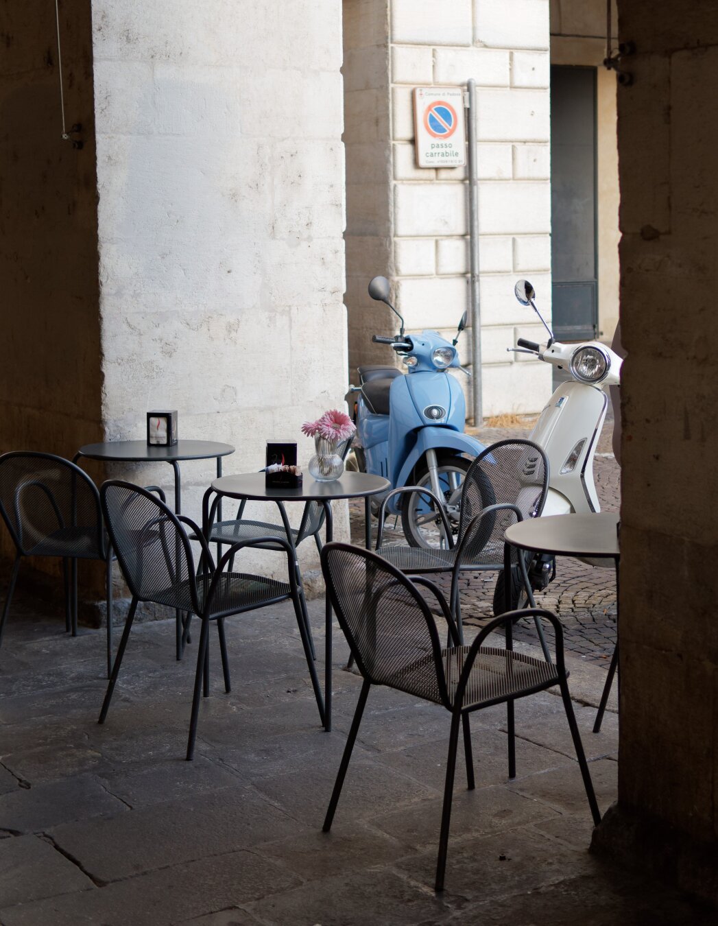 The image captures a charming outdoor café setting in the historic city of Padua, Italy. Nestled under a shaded stone archway, small round tables with minimalist metal chairs are arranged, inviting visitors to pause and enjoy the tranquil atmosphere. A vase with delicate pink flowers and a lit candle on one of the tables adds a touch of warmth and intimacy to the scene. In the background, two vintage-style scooters, one in pastel blue and the other in white, are parked against the stone walls of a narrow street, embodying the timeless charm of Italian city life. The rustic textures of the stone and the gentle interplay of light evoke the elegant and laid-back spirit of Padua.