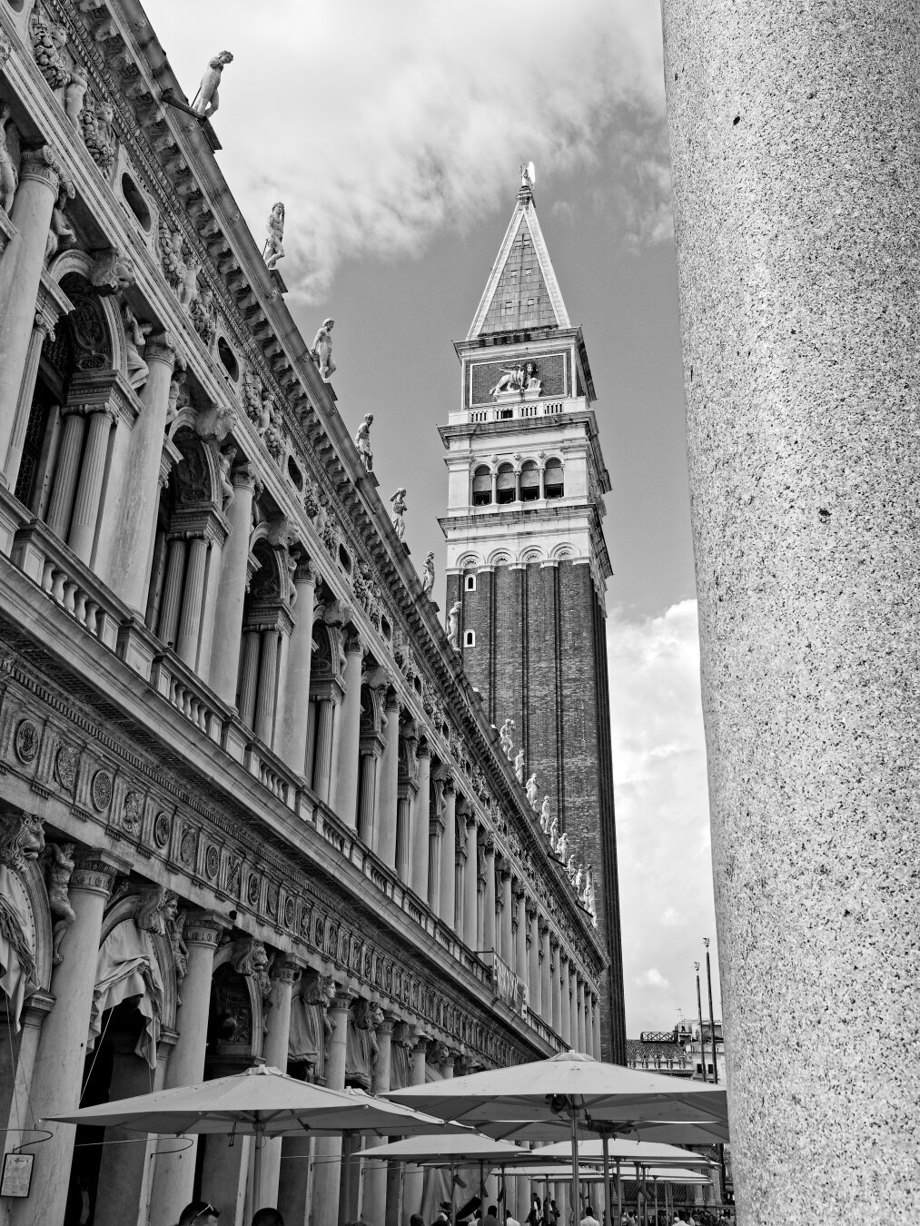 This black-and-white photograph showcases the iconic architecture of St. Mark's Square (Piazza San Marco) in Venice, Italy. The composition highlights the intricate details of the ornate columns and sculptures along the facade of the Procuratie, leading the eye upward to the towering St. Mark's Campanile. The soft clouds in the sky contrast with the sharp lines of the historic buildings, emphasizing their grandeur. In the foreground, café umbrellas add a touch of life and suggest the bustling atmosphere of this famous Venetian landmark.