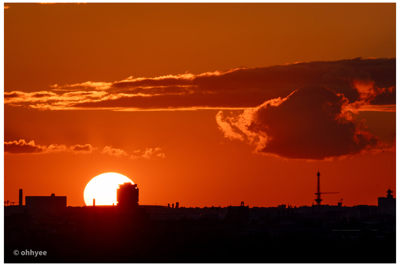 A wide shot of a deep orange and crimson sky at sunset. The bright sun dips near the horizon behind the black silhouette of a city skyline. Thin, dark clouds drift across the vibrant glowing sky.