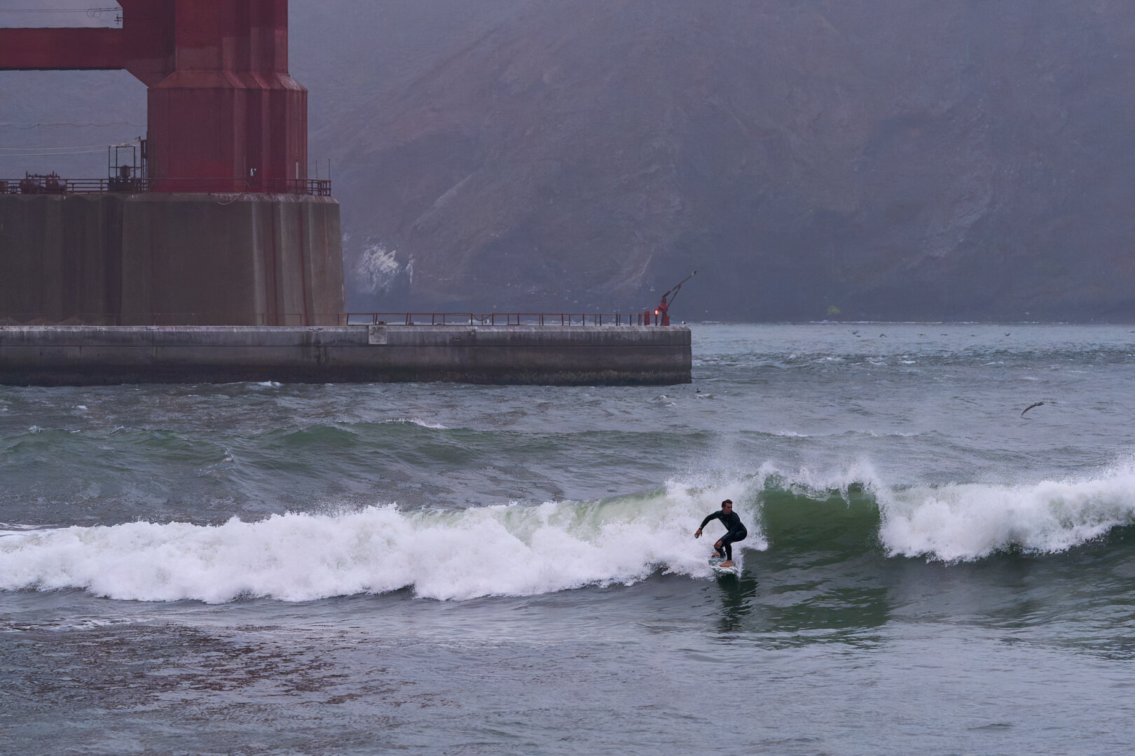 A surfer riding a wave near a tower base of the Golden Gate Bridge. Behind him is the concrete pedestal with the iconic red steel rising from it. It is a gray, gloomy day. You wouldn't know it, but the sun rose a few minutes before this photo.