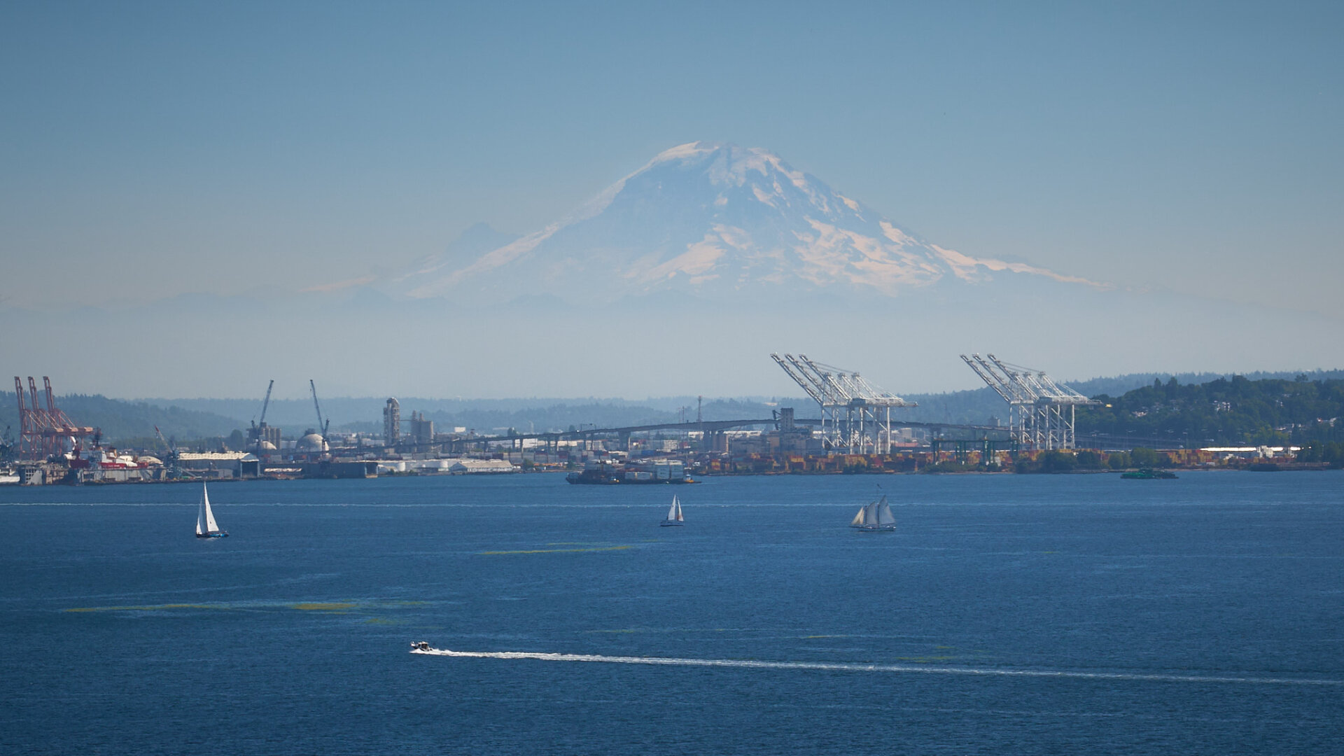 A wide-angle landscape shot depicts a busy harbor with several boats on a deep blue body of water in the foreground. Behind the water, an industrial shoreline is visible, featuring various cranes, shipping containers, and a large bridge. In the far distance, a massive, snow-capped mountain dominates the horizon under a clear, pale blue sky.