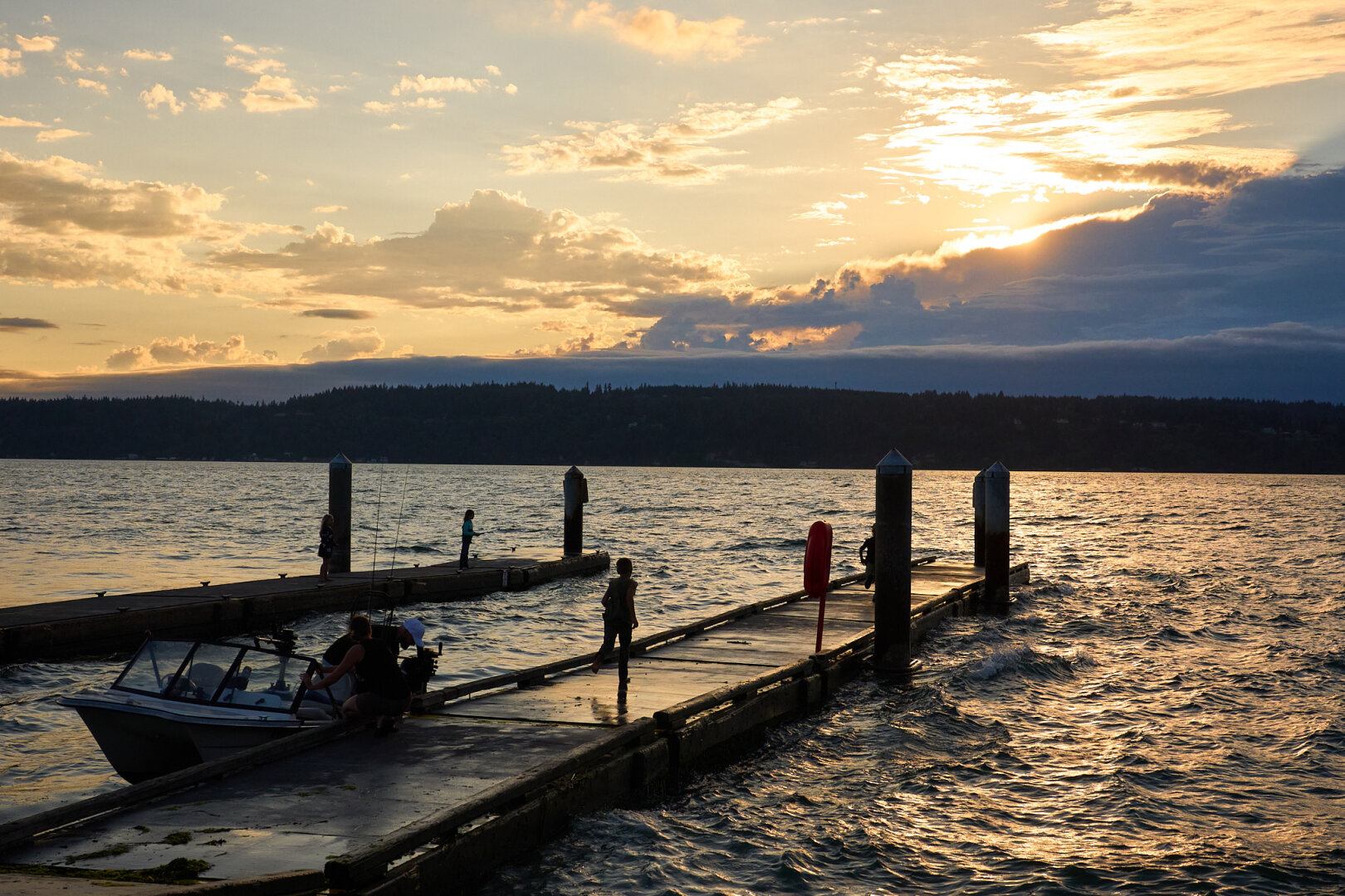 A sunset over a body of water with two wooden piers extending from the shore. Several people are visible on the docks, including one running and others near a small motorboat docked on the left. The sky is filled with clouds illuminated by the setting sun, creating a warm glow that reflects on the surface of the choppy water. In the background, a dark, forested coastline stretches across the horizon.