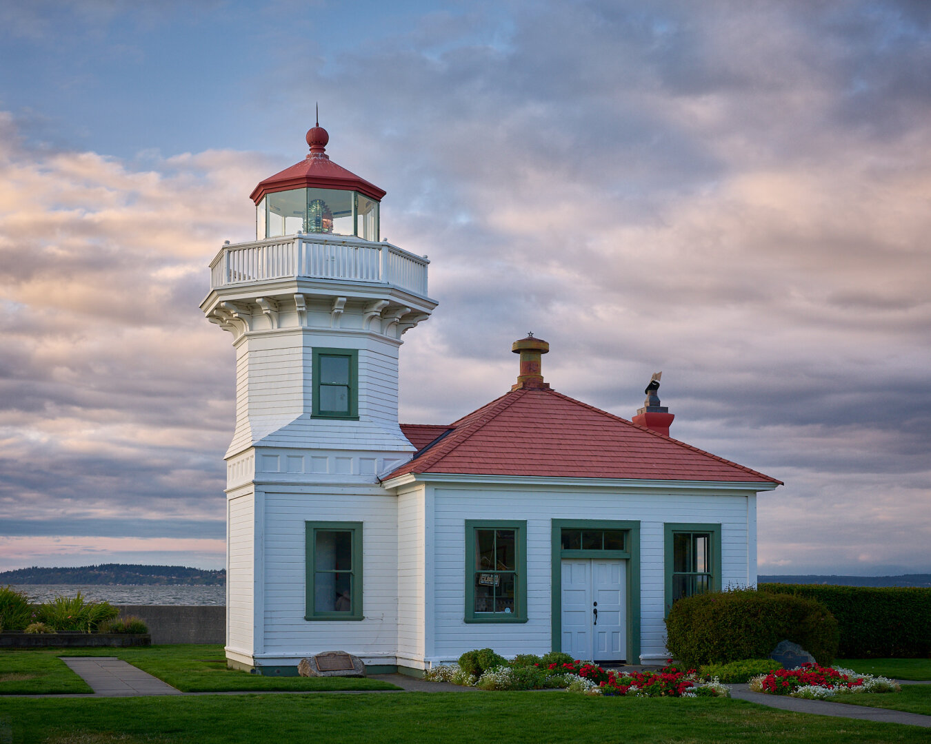 A white, octagonal lighthouse with a red roof, attached to a one-story white building with a matching red roof. The lighthouse and building feature green trim around the windows and doors. They are situated on a green lawn with flower beds and a paved path leading to the entrance. In the background, a body of water is visible under a cloudy, sunset-lit sky.
