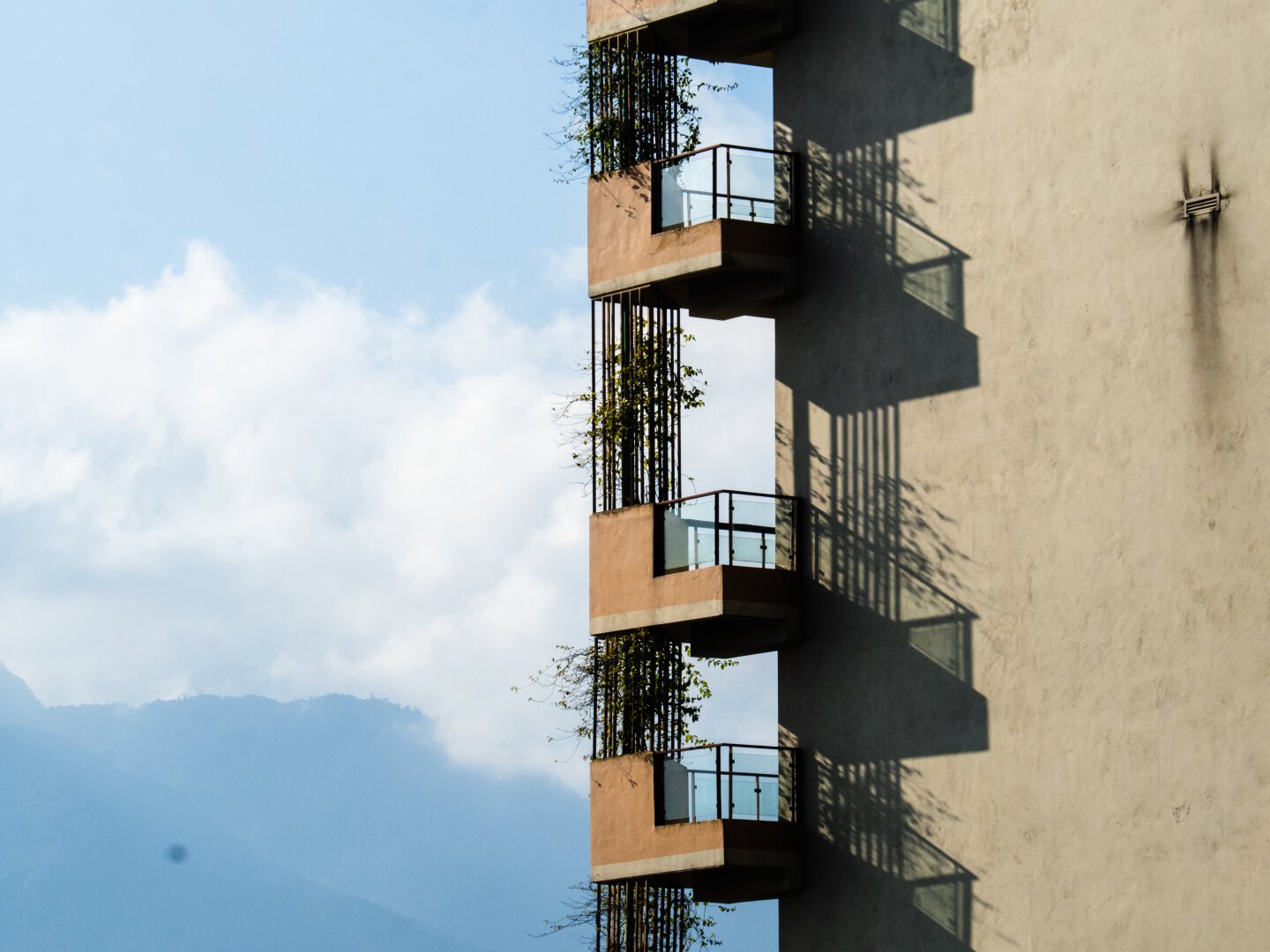 A hazy blue sky and mountains on the left is contrasted sharply by the edge of an apartment building featuring shadows from the balconies...