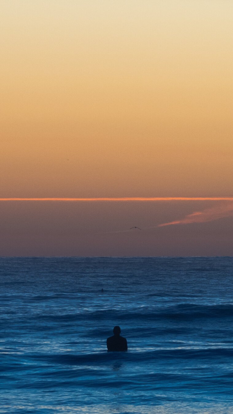 A surfer is silhouetted floating in the ocean at sunrise