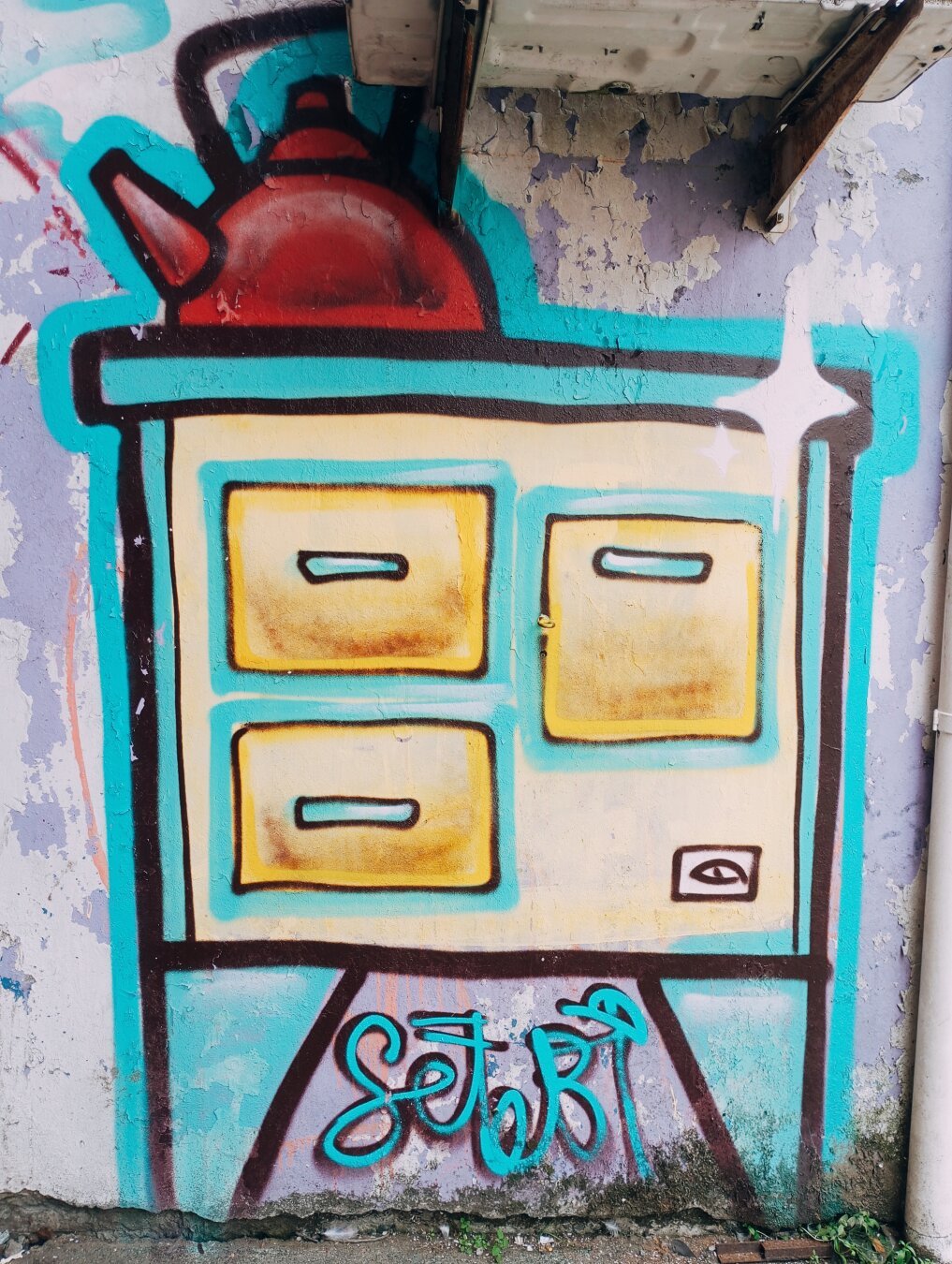 grafiti en una pared descascarada muestra una tetera roja sobre un mueble de estilo antiguo con cuatro cajones amarillos. El mueble está pintado en tonos turquesa y beige, con líneas negras que definen sus bordes y cajones. Debajo del mueble, la firma "Setebi" está escrita en un estilo cursivo turquesa. La pared muestra signos de deterioro y desgaste.