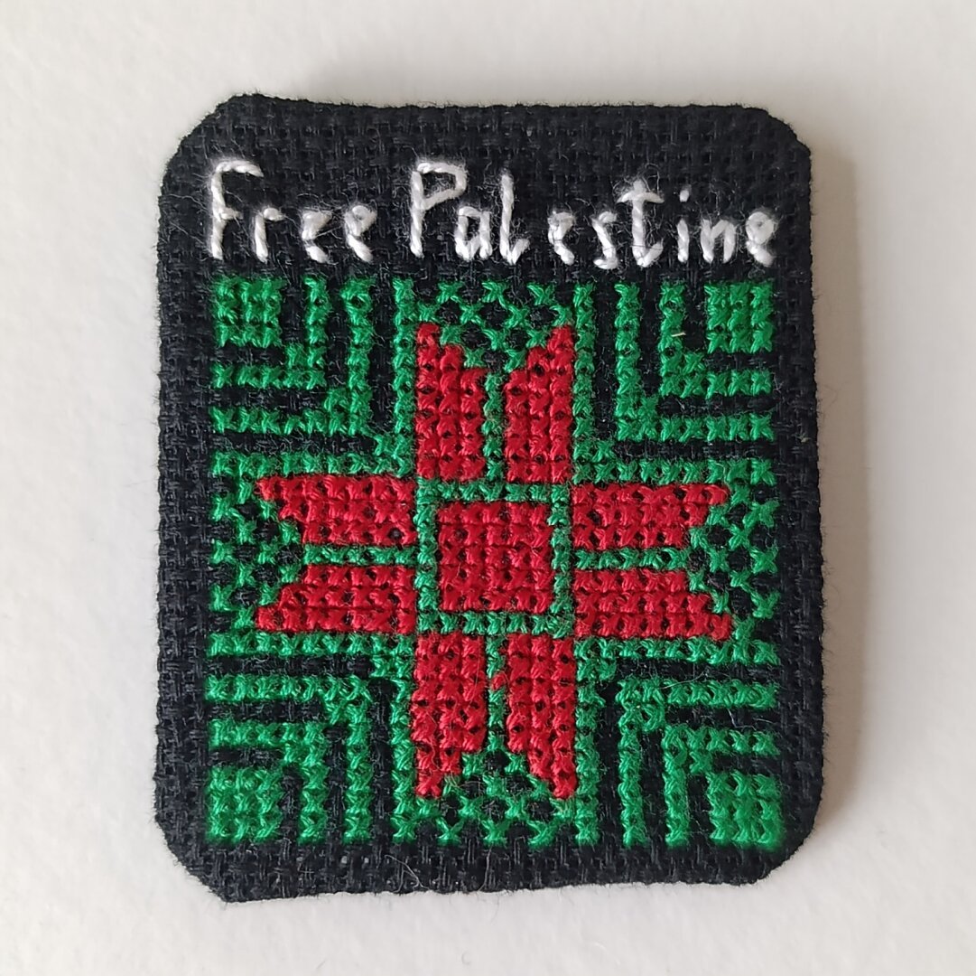 A crosstitch pin with red star, green pattern and white words "Free Palestine" at the top.