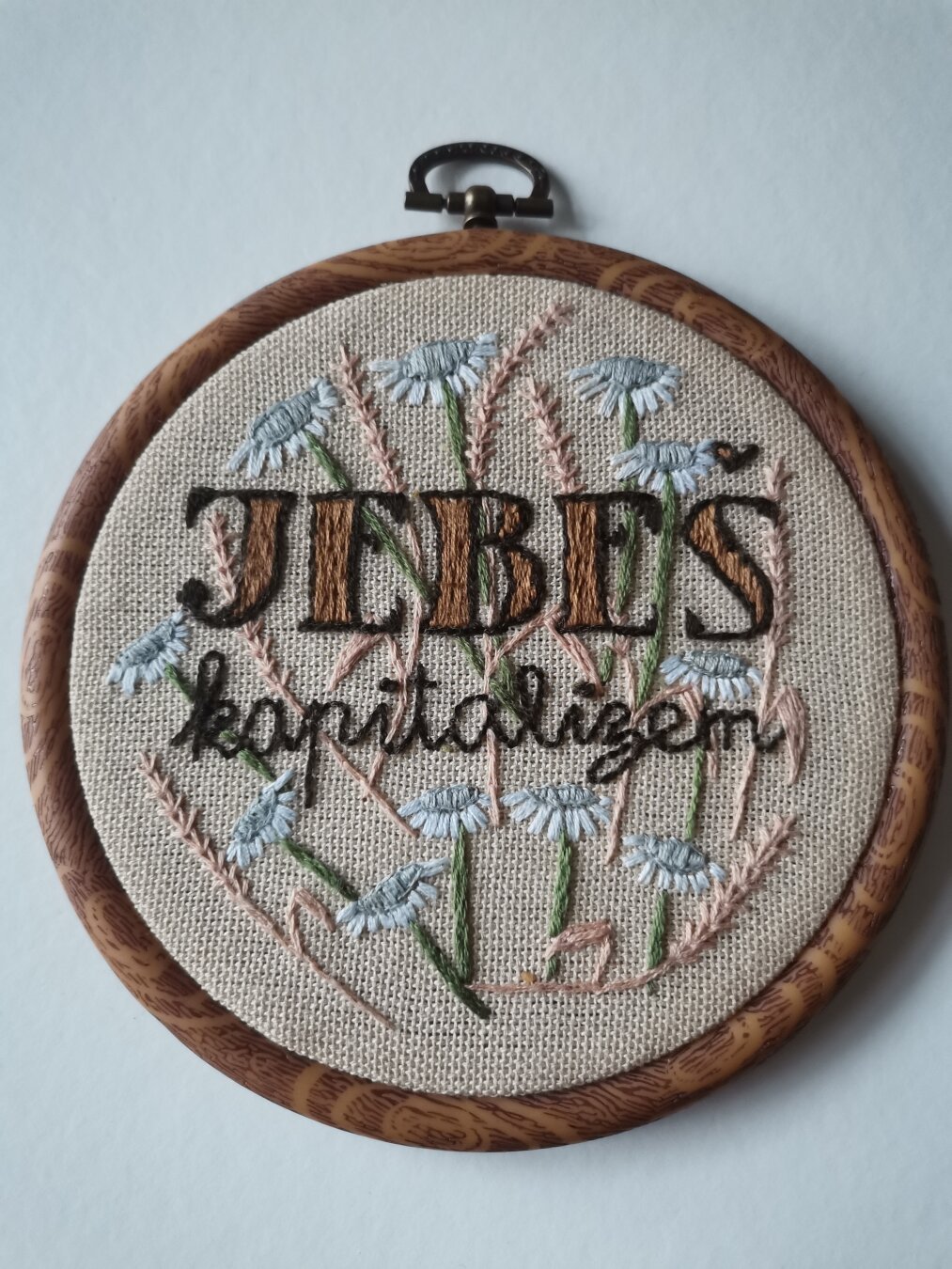 Okrogel obroč za vezenje z izvezenim napisom "jebeš kapitalizem", okrašenim z vezeninami cvetja in klasja.