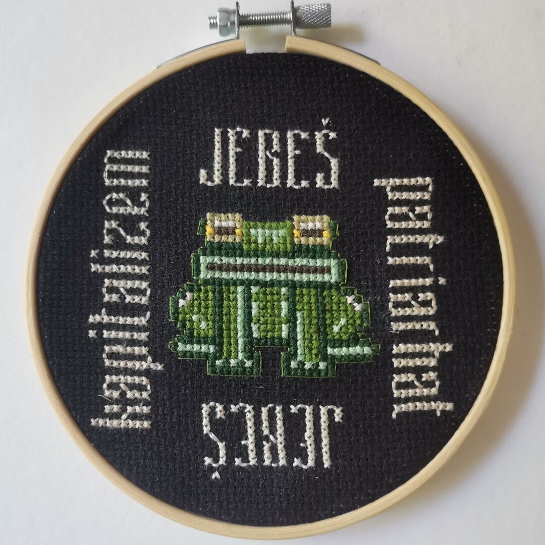 Obroč za vezenje z vezenino v crosstitch tehniki. Na sredini zelena žaba, okrog nje napisano: nad in pod njo "jebeš", levo "kapitalizem", desno "patriarhat".