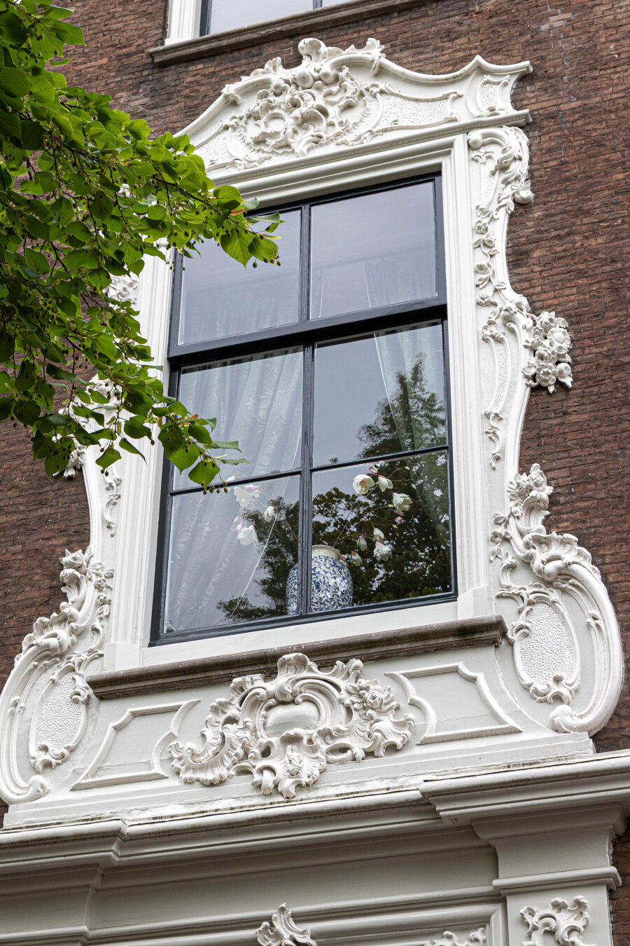 A window of a historic building in Delft, surrounded by white plasterwork, full of curls and flowers. Photographed from below. In front of the window is a Delft blue vase with white flowers. The sky and a tree are reflected in the window. A bit of green from that tree is visible in the upper left of the photo. A window of a historic building in Delft, surrounded by white plasterwork, full of curls and flowers. Photographed from below. In front of the window is a Delft blue vase with white flowers. The sky and a tree are reflected in the window. A bit of green from that tree is visible in the upper left of the photo.