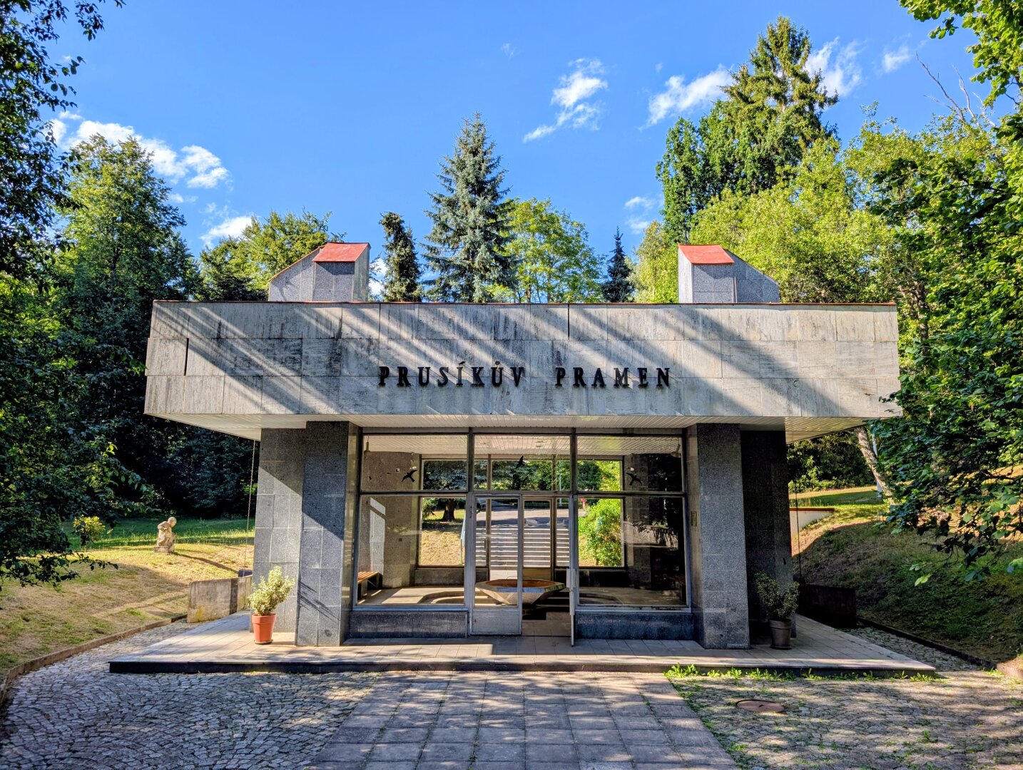 The photo shows a retro-modern pavilion with the sign "PRUSÍKŮV PRAMEN" on its facade, surrounded by lush trees and a well-maintained park under a bright blue sky. Large glass windows and clean architectural lines give the building a simple appearance.