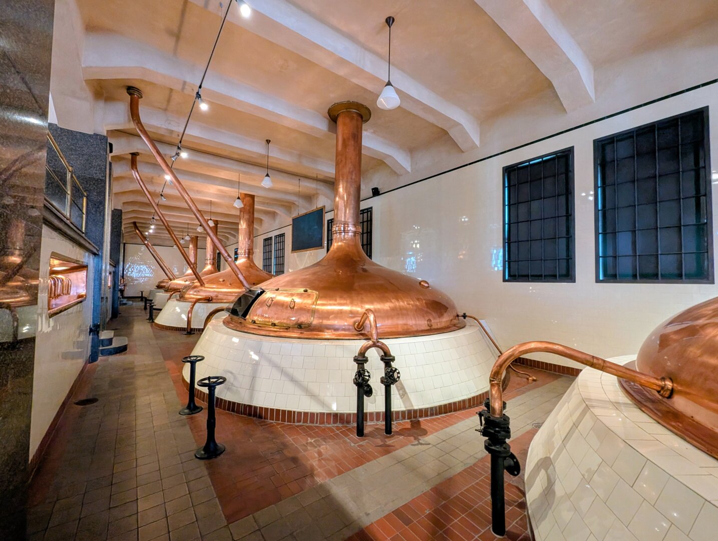 The photo shows a row of large copper brewing kettles inside a clean, historic brewery, with white tiled bases and polished copper pipes stretching across the room. The high arched ceiling, industrial lighting, and symmetrical layout create a spacious, well-maintained atmosphere that highlights traditional brewing equipment.