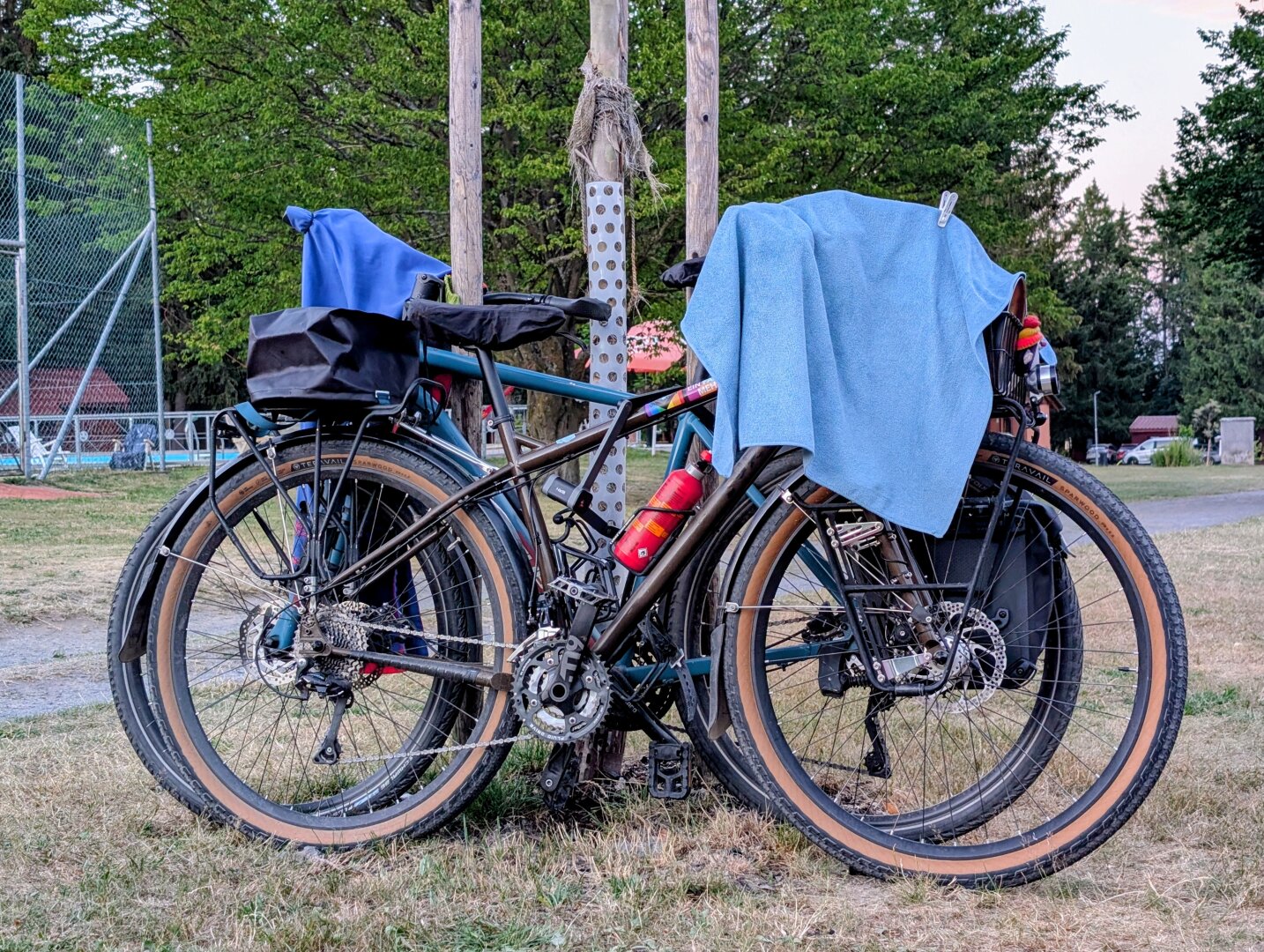 The photo shows two touring bicycles parked side by side against a wooden post, with towels draped over them. Surrounded by trees and a grassy area, the scene suggests the cyclists are taking a break during a long outdoor ride.
