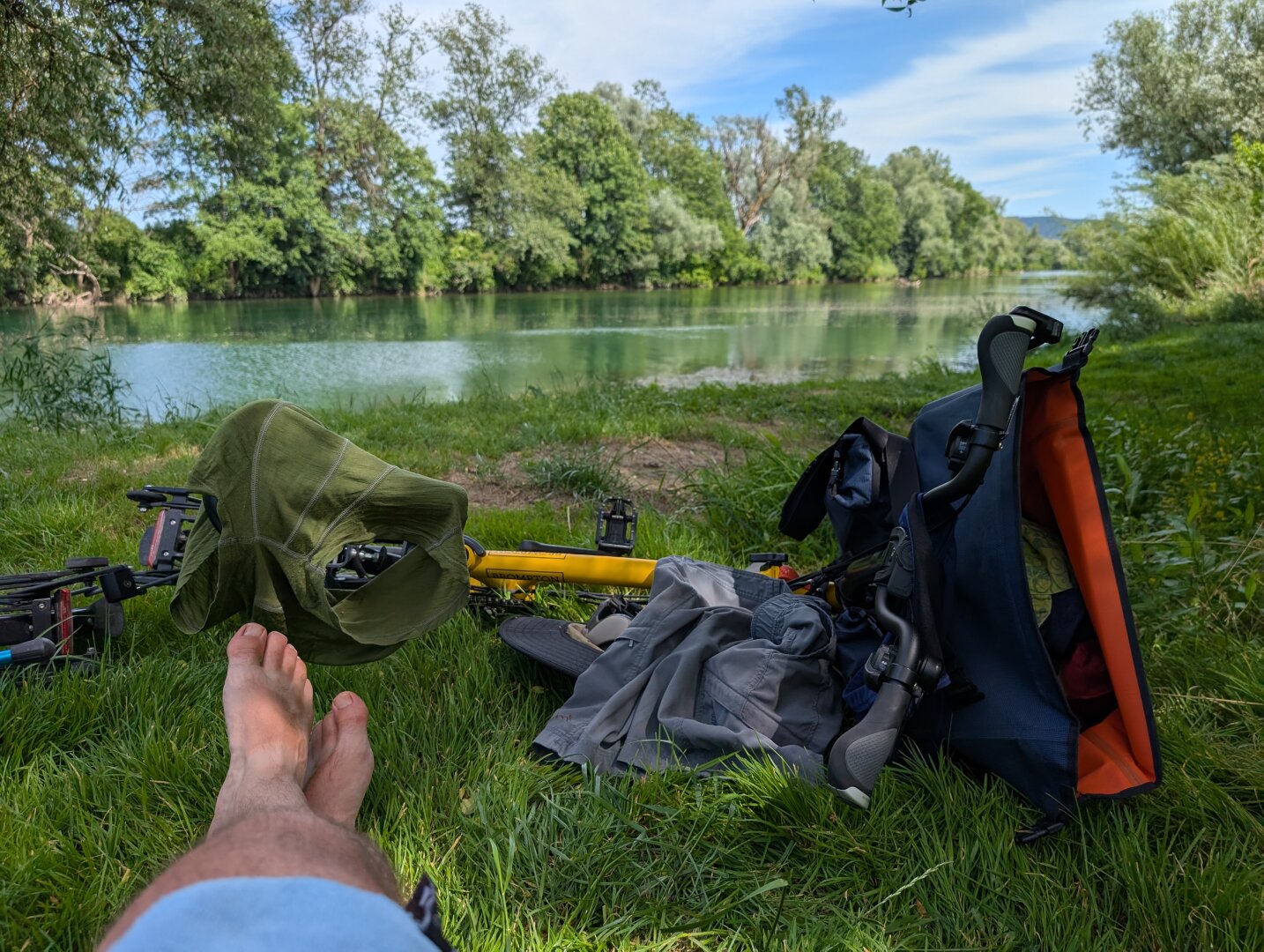 In foremost part there is a folding bicycle with a front bag attached, laying on its side in grass. There are bare male feet visible also, from someone laying in the grass. In the background there is a river with trees growing on its banks.