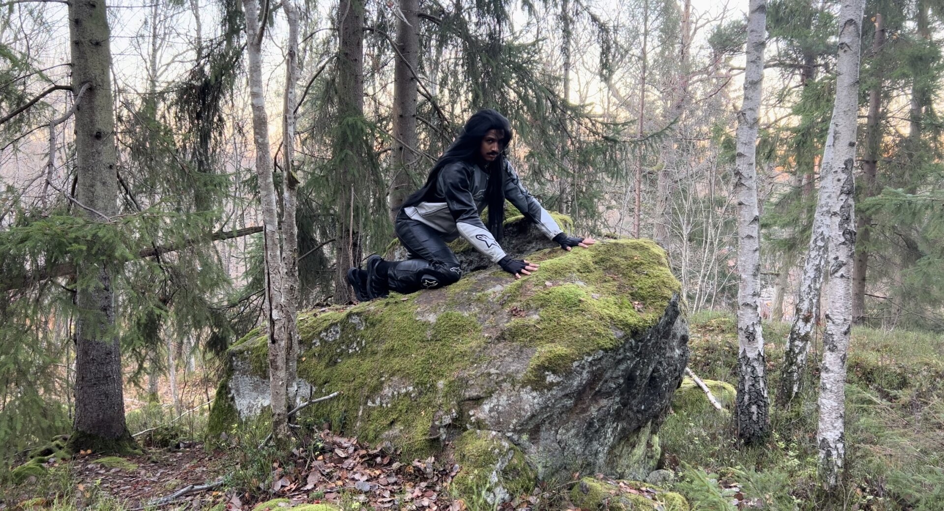 Adam on a rock in the forest, wearing black and grey leather motorcycle gear, and a long black wig.