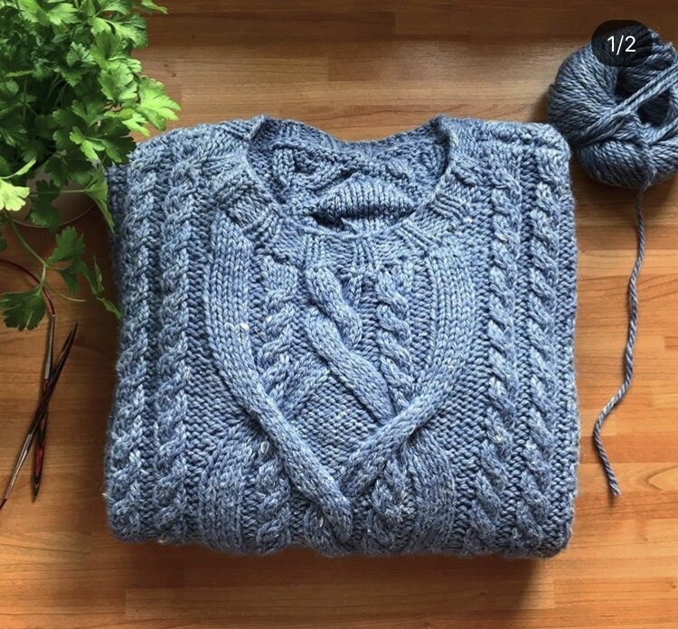 A picture of a folded-up knitted jumper. The jumper is knitted with a chunky yarn and features various cable patterns. Next to the jumper is a partially used ball of a yarn and a small plant.